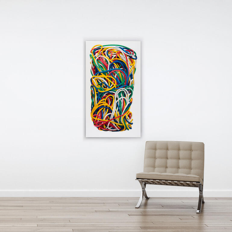 Buntes Gummiband-Kunstwerk hängt an einer weißen Wand über einem Stuhl.
