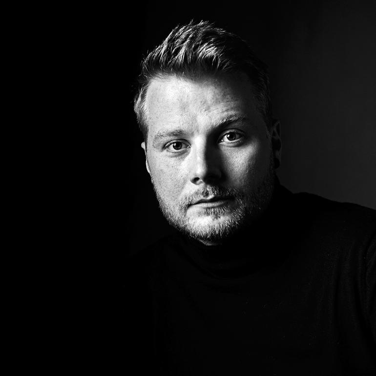 David Tollmann Porträt eines Mannes mit Bart, schwarzer Rollkragenpullover vor dunklem Hintergrund.