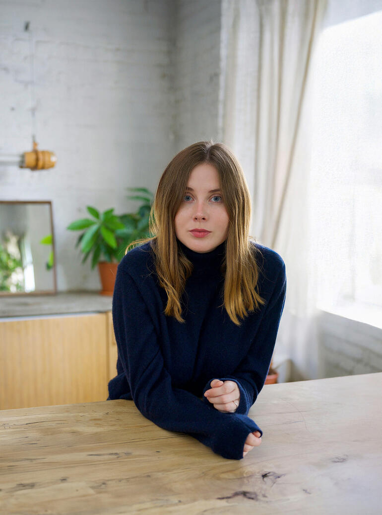 Charlotte Rother Junge Frau in einem dunkelblauen Pullover sitzt am Tisch und blickt direkt in die Kamera.