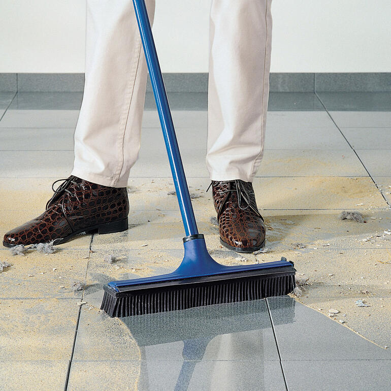 Person fegt mit einem blauen Besen, Sand und Dreck auf dem Boden.